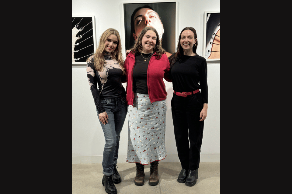 Three women standing together in an art gallery in front of a wall of photographs of women