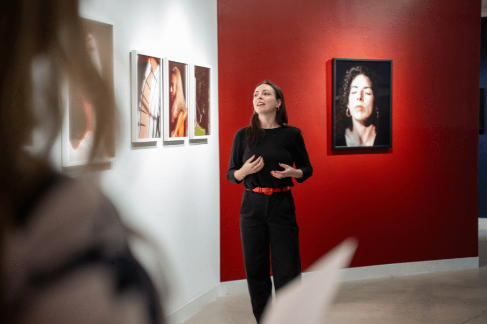 A woman standing in front of a wall of photographs of women speaking to people at an art gallery